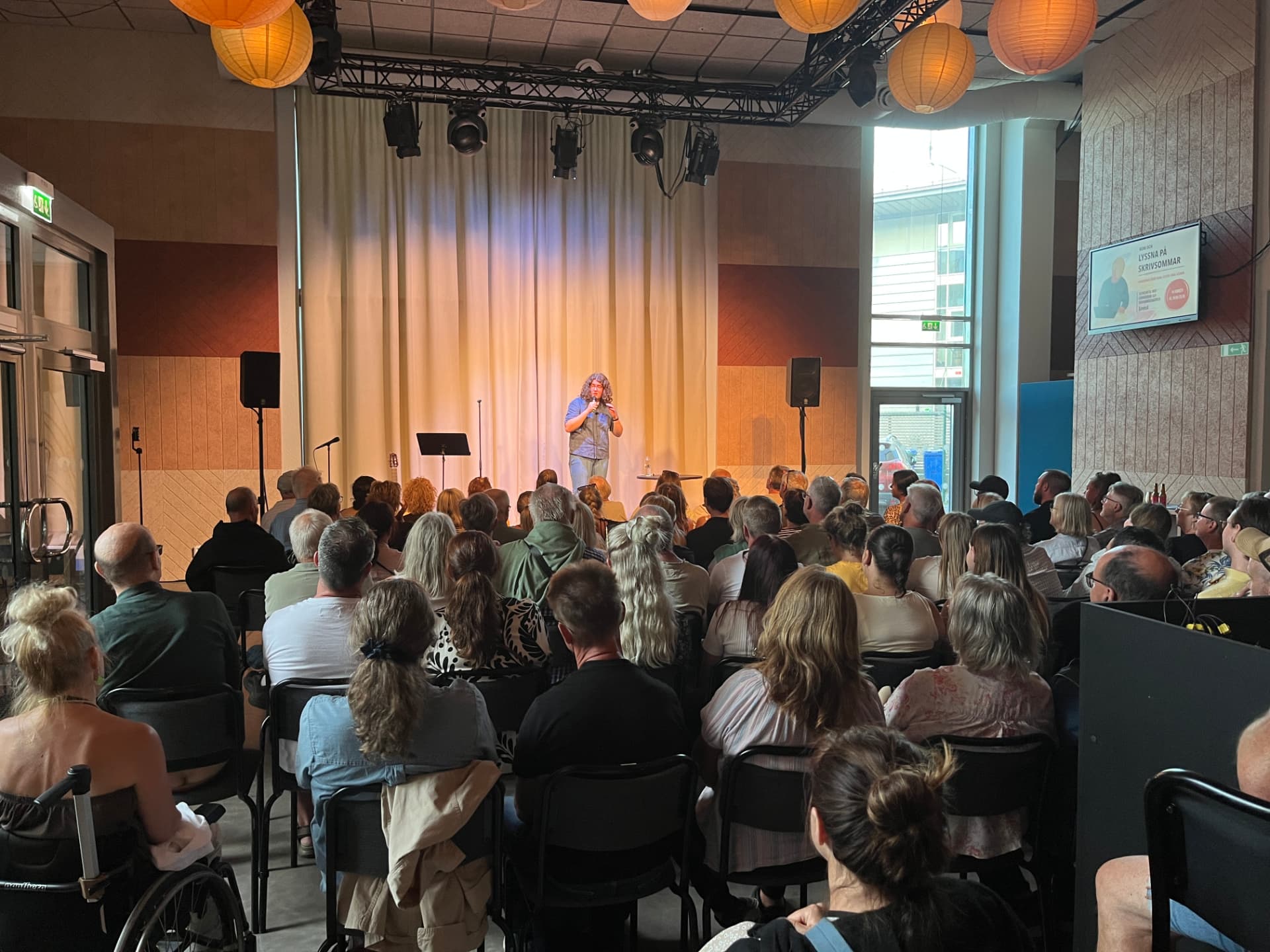 Komiker med mikrofon på scen framför fullsatt publik i Byteaterns foajé i Kalmar