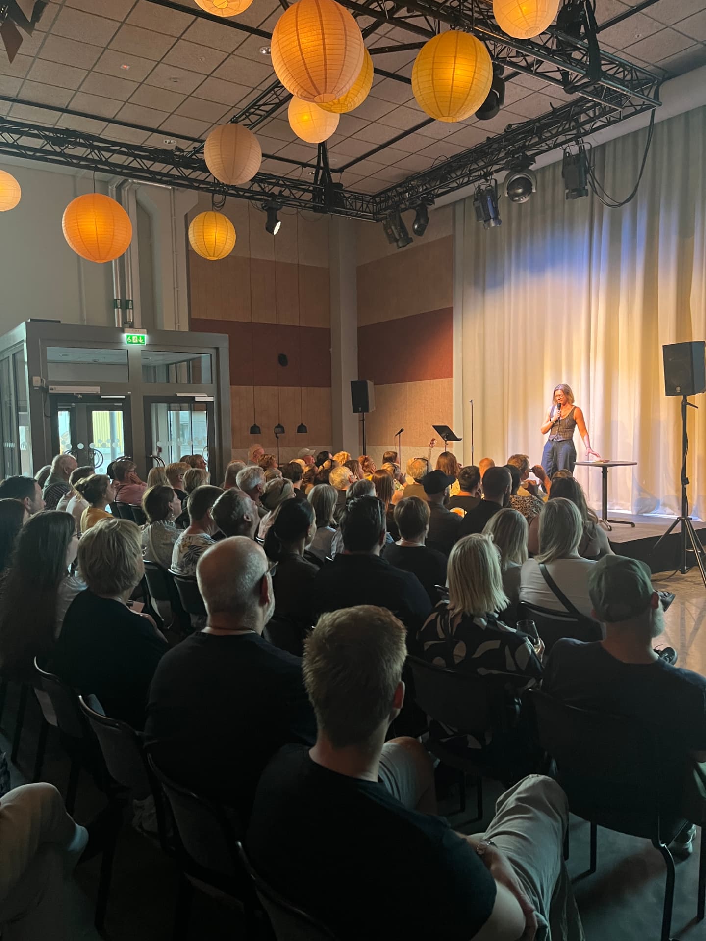 Publikvy mot scenen med lyktor i taket under en stand-up-kväll