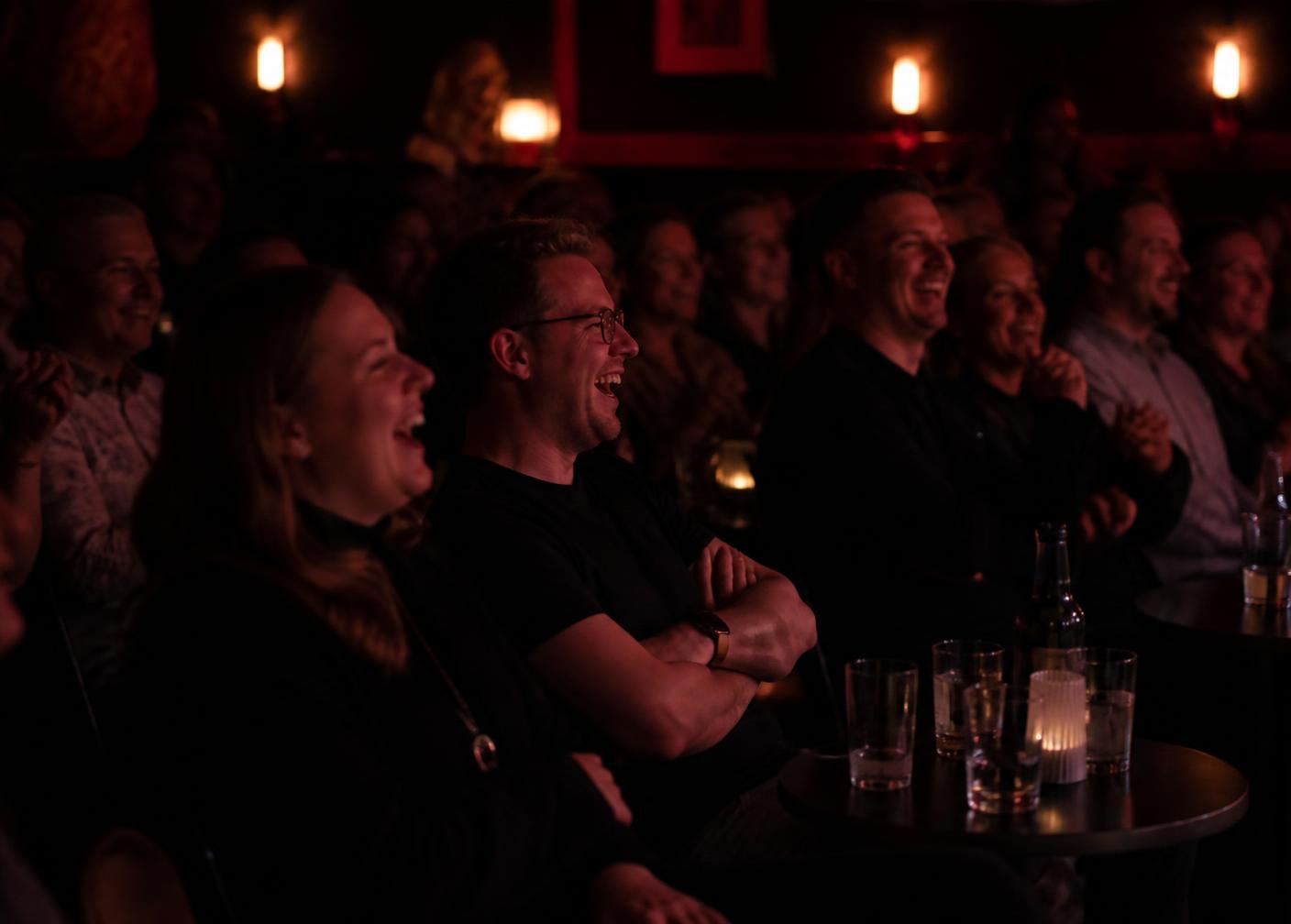 Skrattande publik vid små bord i en mörk comedyklubb